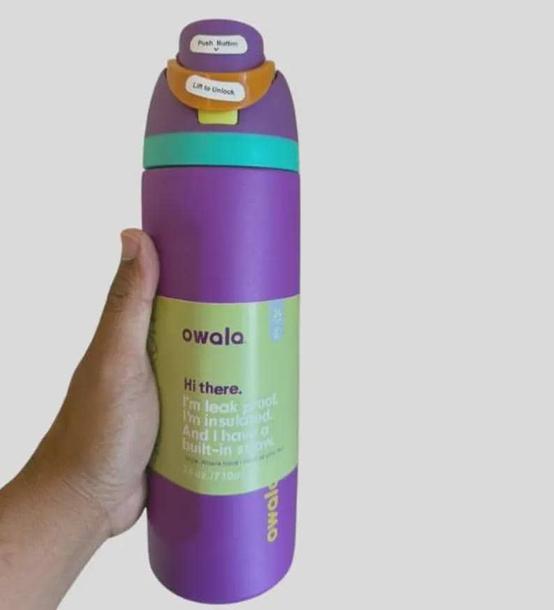Op de afbeelding: Een paarse Owala-waterfles met een turquoise band, een oranje vergrendelingsmechanisme en een paars deksel. Het label leest: Hi there. I'm leak proof. I'm insulated. And I have a built-in straw. 24 oz / 710 ml. Het deksel bevat de tekst Push Button en Lift to Unlock.