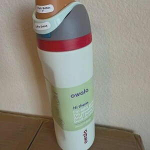 Op de afbeelding: Een witte Owala-waterfles met een meerkleurig deksel met een bruine drukknop, een lichtblauw vergrendelingsmechanisme en een grijze dop. De fles heeft een groen label met de tekst Hi there. I'm leak proof. I'm insulated. And I have a built-in straw. 24 oz / 710ml.