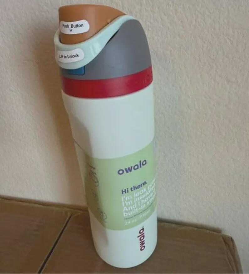 Op de afbeelding: Een witte Owala-waterfles met een meerkleurig deksel met een bruine drukknop, een lichtblauw vergrendelingsmechanisme en een grijze dop. De fles heeft een groen label met de tekst Hi there. I'm leak proof. I'm insulated. And I have a built-in straw. 24 oz / 710ml.