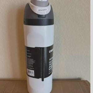 Op de afbeelding: Een witte waterfles met een donkergrijze dop voorzien van een drukknop en een ontgrendelingsmechanisme. Een zwart productlabel is om het midden gewikkeld, met witte tekst en een streepjescode. De fles heeft een inhoud van 710 ml op het label.