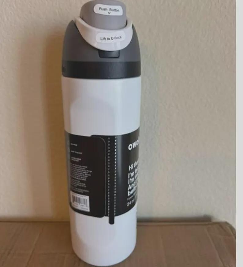 Op de afbeelding: Een witte waterfles met een donkergrijze dop voorzien van een drukknop en een ontgrendelingsmechanisme. Een zwart productlabel is om het midden gewikkeld, met witte tekst en een streepjescode. De fles heeft een inhoud van 710 ml op het label.