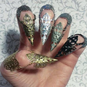 Könnte beinhalten: Satz von sechs Fingerringen mit einem spitzen, klauenartigen Design. Die Ringe sind aus Metall und haben verschiedene Oberflächen, darunter Silber, Gold und Schwarz.