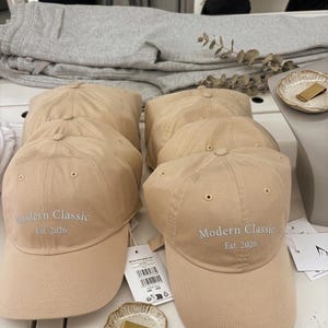 May include: A beige baseball cap featuring the embroidered text Modern Classic Est. 2026 on the front panel. The cap has a structured crown, a curved brim, and stitched eyelets for ventilation. A white product tag is attached to the side of the cap.