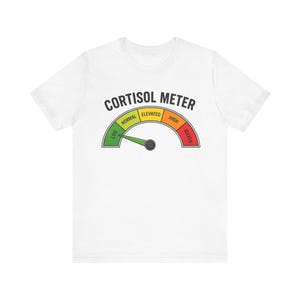May include: A white short-sleeve t-shirt featuring a graphic of a gauge labeled CORTISOL METER. The gauge has sections colored green, yellow, orange, and red, with labels for LOW, NORMAL, ELEVATED, HIGH, and MAXED, and a green needle pointing to LOW.