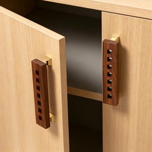 May include: Two rectangular cabinet handles made of dark brown wood with a vertical row of square cutouts. Each handle is attached to a light oak wood cabinet door with polished gold-tone metal mounting brackets at the top and bottom ends.
