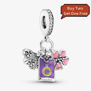 May include: A silver dangle charm featuring a bail encrusted with clear stones. Three pendants hang from the bail: a silver fan, a rectangular purple enamel tag with a yellow ring icon and the text PANDORA, and a pink five-petaled flower.