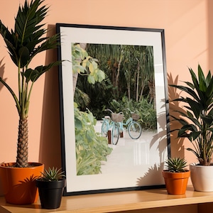 May include: A framed vertical wall art print featuring two light blue bicycles with wicker baskets parked on a sandy path surrounded by lush green tropical foliage. The print is enclosed in a thin black frame and displayed on a wooden surface.