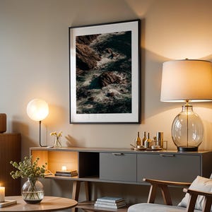 Puede incluir: Una lámina de arte mural vertical enmarcada que muestra acantilados rocosos oscuros y olas rompiendo, sobre un aparador gris. La escena incluye una lámpara de mesa de cristal con pantalla blanca, un jarrón de cristal redondo y una vela.