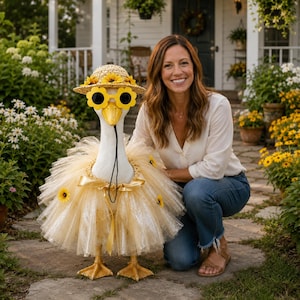 May include: A white goose statue wearing a straw hat decorated with sunflowers, yellow flower-shaped sunglasses, and a layered gold tulle tutu skirt adorned with small sunflowers. The goose has a bright orange beak and stands on orange feet.