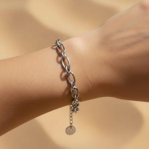 Könnte beinhalten: Ein silberfarbenes Gliederarmband mit länglichen, ovalen Kettengliedern. Das Armband verfügt über eine kurze Verlängerungskette, die in einem kleinen, runden Metallanhänger endet. Die Glieder sind poliert und in einem gleichmäßigen Muster verbunden.