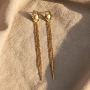 Könnte beinhalten: Ein Paar goldfarbene Ohrhänger mit einem sonnenförmigen Stecker und einem zentralen, weißen, ovalen Cabochon. Jeder Ohrring hat zwei lange, schmale, goldfarbene Ketten, die vertikal vom Stecker herabhängen und eine zarte, längliche Form bilden.