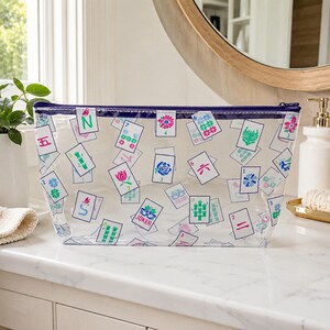 May include: A clear rectangular zippered pouch featuring a repeating pattern of colorful mahjong tile illustrations. The design includes various tiles with numbers, bamboo symbols, flowers, and a Joker tile, all printed in vibrant shades of blue, pink, and green.