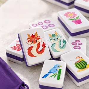K&ouml;nnte beinhalten: Eine Sammlung rechteckiger Mahjong-Spielsteine mit wei&szlig;en Oberseiten und violetten Unterseiten. Die Steine zeigen farbenfrohe Illustrationen, darunter einen roten Drachen, einen gr&uuml;nen Drachen, einen blauen Vogel auf einem Bambuszweig, ein Wassermelonengetr&auml;nk, eine rosa Blume und violette Blumenmuster.
