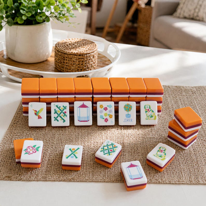 K&ouml;nnte beinhalten: Ein Set rechteckiger Mahjong-Spielsteine mit orangefarbenen R&uuml;ckseiten und wei&szlig;en Vorderseiten, die bunte Illustrationen von Blumen, einem Kolibri, einem Pavillon, einem Hund und einem Formschnitt zeigen. Einige Steine sind gestapelt.