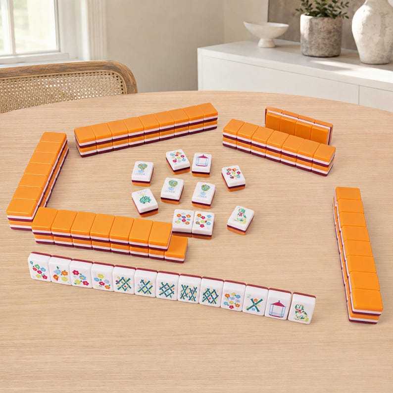 K&ouml;nnte beinhalten: Ein Mahjong-Spielset mit orangefarbenen R&uuml;ckseiten und wei&szlig;en Vorderseiten, die mit bunten Blumen-, geometrischen und architektonischen Illustrationen versehen sind. Die Spielsteine sind in Stapeln und einer Reihe angeordnet und zeigen verschiedene Muster.