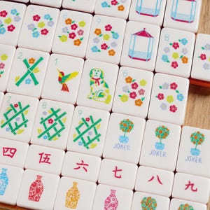 K&ouml;nnte beinhalten: Eine Sammlung wei&szlig;er, rechteckiger Mahjong-Spielsteine mit farbenfrohen Illustrationen von Blumen, einem Kolibri, einem Hund, Pavillons, Bambusmustern, als JOKER beschrifteten Topfb&auml;umen und verschiedenen Vasen sowie roten chinesischen Schriftzeichen.