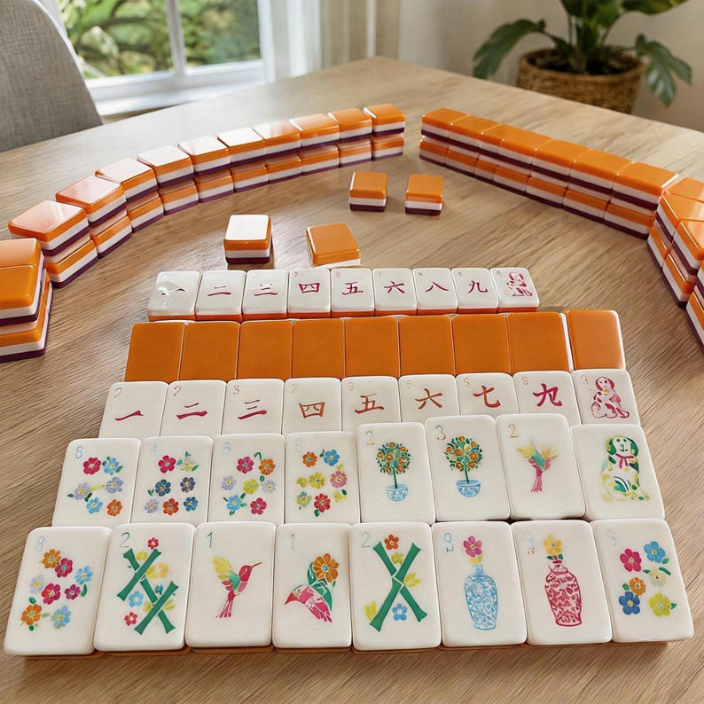 K&ouml;nnte beinhalten: Ein Mahjong-Spiel mit rechteckigen Spielsteinen, die orangefarbene R&uuml;ckseiten und wei&szlig;e Vorderseiten haben. Die Steine zeigen verschiedene Motive wie chinesische Ziffern, bunte Blumenmuster, Topfb&auml;ume, einen Kolibri, Vasen und einen Hund.