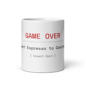 Può includere: Una tazza in ceramica bianca con il testo rosso GAME OVER in alto, seguito da una linea orizzontale e dal testo nero Insert Espresso to Continue, con il testo [ Insert Coin ] stampato in nero nella parte inferiore della superficie.