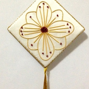 May include: A white square ornament with a gold trim and a gold tassel. The ornament features a gold embroidered flower with a red center.