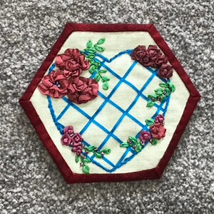 May include: A hexagonal fabric patch with a red border and a white background. The center of the patch features a blue grid design in the shape of a heart. The heart is surrounded by red ribbon roses and green leaves.