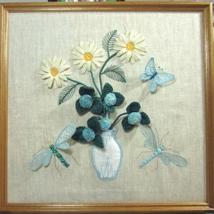 May include: A framed embroidery depicting a white vase with a bouquet of white daisies and blue butterflies and dragonflies. The embroidery is on a beige linen background.