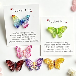 Puede incluir: Una colección de dijes de resina con forma de mariposa colorida. Dos dijes están sujetos a tarjetas blancas con el texto Pocket Hug y un poema. Otras mariposas sueltas en rojo, rosa, morado y amarillo están dispuestas debajo de las tarjetas.