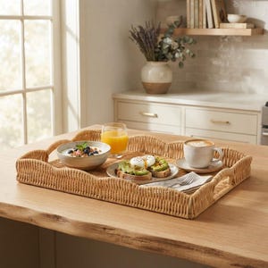Puede incluir: Una bandeja de servicio rectangular de ratán trenzado con bordes festoneados y asas integradas. La bandeja contiene un cuenco de cerámica con yogur y arándanos, un plato con tostadas de aguacate y huevo poché, un vaso de zumo de naranja y una taza de café.