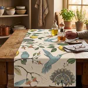 Op de afbeelding: Een rechthoekige tafelloper met een crèmekleurige achtergrond en een botanische print van blauwe kolibries, groene bladeren, oranje bloemen en gele citroenen. Het ontwerp bevat gedetailleerde illustraties van vogels in de vlucht en op takken.