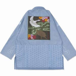 Op de afbeelding: Een lichtblauw jasje met een grote vierkante patch op de rug met een geborduurde witte kraanvogel, een bruine schildpad en kleurrijke bloemen. Het onderste deel heeft een wit golfpatroon en de mouwen zijn voorzien van witte wolkenmotieven.
