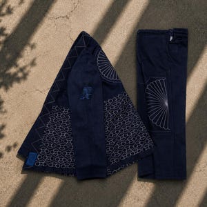 May include: A dark navy blue jacket and matching trousers set. The jacket features a zigzag stitched collar, a blue embroidered AP logo on the sleeve, and a white floral pattern on the lower panels. The trousers have a white fan-like graphic.