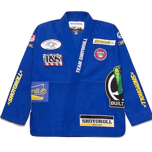 Puede incluir: Una chaqueta de gi para artes marciales en color azul real con mangas largas y cuello estructurado. La prenda está adornada con varios parches bordados, incluyendo un gráfico de cobra y el texto SHOYOROLL, TEAM SHOYOROLL, P&S y CRUSH.