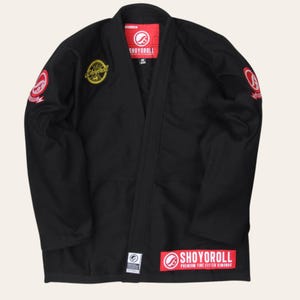 Könnte beinhalten: Eine schwarze Kampfkunst-Gi-Jacke mit einem rot-weißen Shoyoroll-Logo am Innenkragen, einem gelben runden Aufnäher auf der rechten Brust und rot-weißen runden Aufnähern an beiden Ärmeln. Ein roter rechteckiger Aufnäher befindet sich auf der unteren Vorderseite.
