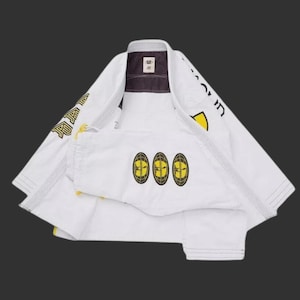 May include: A white martial arts gi jacket featuring long sleeves with black and yellow graphic text. The chest area displays three circular yellow and black globe logos. The interior collar is dark gray with a small white brand label.