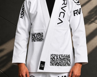May include: A white martial arts gi featuring black RVCA branding and text on the chest and sleeves. The garment includes rectangular patches with intricate black graffiti-style lettering on the front and a black label near the hem.