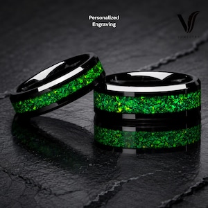 Puede incluir: Dos anillos de metal negro con una incrustación central de piedra verde triturada. Los anillos tienen un acabado pulido y un diseño de banda lisa. El texto Personalized Engraving y el logotipo de Vowistry son visibles en la parte superior.