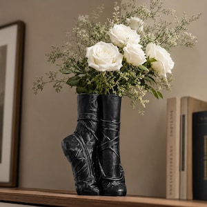 May include: A black vase shaped like a pair of ballet pointe shoes, featuring detailed ribbons and laces. The vase is filled with a bouquet of white roses and delicate green filler flowers, creating a contrast between the dark footwear and blooms.