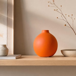 Puede incluir: Un jarrón naranja de forma esférica con un acabado mate liso y una abertura circular estrecha en la parte superior. El jarrón está colocado sobre una superficie de madera clara, destacando como un objeto vibrante y de color sólido sobre un fondo neutro.