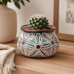May include: A small, round ceramic planter featuring a textured surface with a green and beige speckled finish. The pot is decorated with white-outlined flower motifs that have dark red centers. A small, spiky green cactus sits inside the planter.