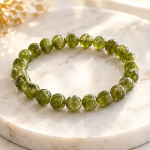 Pode incluir: Uma pulseira elástica composta por contas redondas de pedra preciosa em verde-oliva polido. As contas exibem uma qualidade translúcida com variações internas subtis de cor e textura, dispostas num círculo contínuo sobre uma superfície.