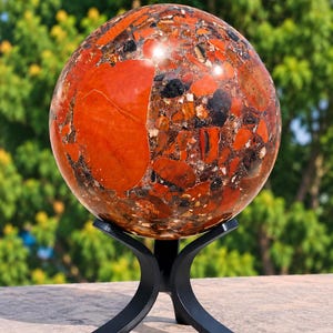 Puede incluir: Una esfera de piedra pulida con un patrón brechoide de fragmentos minerales rojos, marrones y negros. La esfera mide 110 mm de diámetro y pesa 1,79 kg. Se exhibe sobre un soporte de metal negro de tres patas.