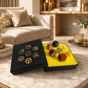 Op de afbeelding: Een zwarte doos met gouden geometrische symbolen op het deksel, met zeven verschillende veelvlakken, waaronder een donkerblauwe bol, een houten kubus en diverse doorschijnende geometrische vormen op een felgele schuimrubberen inzetstuk.