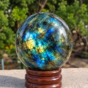 Può includere: Una sfera di labradorite lucida che mostra riflessi iridescenti blu e gialli sulla sua superficie scura e fratturata, poggiata su una base circolare in legno marrone scuro. La pietra presenta una complessa rete di venature interne.