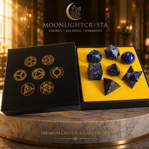 Op de afbeelding: Een zwarte doos met zeven geometrische kristalvormen van blauwe lapis lazuli op een felgele inzet. Het deksel bevat gouden geometrische lijntekeningen, de tekst MOONLIGHTCRYSTA en de woorden ENERGY | BALANCE | HARMONY.