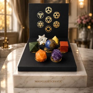 Op de afbeelding: Een zwarte displaydoos met gouden geometrische symbolen en de tekst MOONLIGHTCRYSTA, met zeven verschillende veelhoekige kristallen in diverse kleuren zoals transparant, groen, blauw, oranje, paars, geel en rood, geplaatst op een marmeren voet.