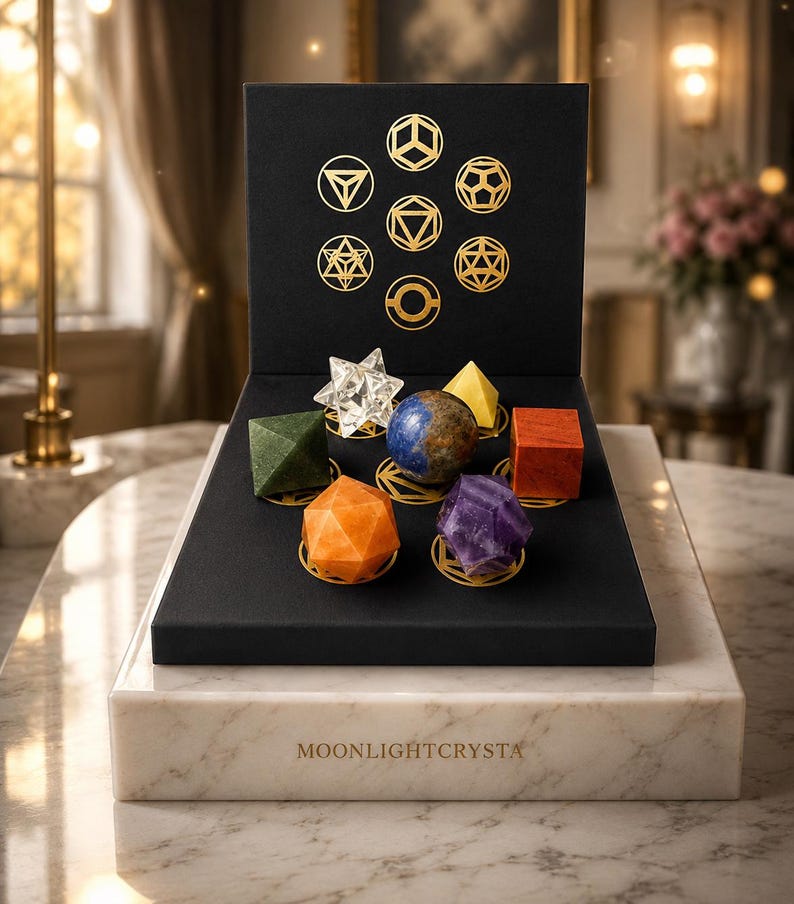 Op de afbeelding: Een zwarte displaydoos met gouden geometrische symbolen en de tekst MOONLIGHTCRYSTA, met zeven verschillende veelhoekige kristallen in diverse kleuren zoals transparant, groen, blauw, oranje, paars, geel en rood, geplaatst op een marmeren voet.
