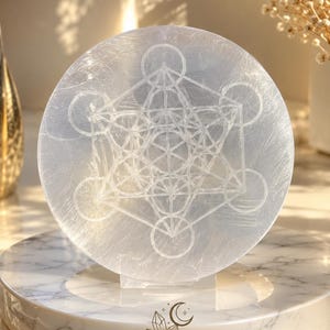 May include: A circular, translucent white selenite crystal slab featuring an etched Metatron's Cube geometric pattern. The crystal has a fibrous, shimmering texture and is positioned upright on a circular white marble base with the text MOONLIGHT CRYSTAL.