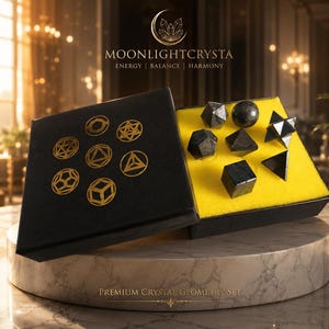 Op de afbeelding: Een zwarte doos met zeven donkere, gepolijste geometrische kristalvormen in felgeel schuim. Het deksel bevat goudkleurige lijntekeningen van diverse veelvlakken en de tekst MOONLIGHTCRYSTA ENERGY | BALANCE | HARMONY.
