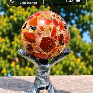 Peut inclure: Une pierre sphérique polie avec un motif mosaïque multicolore composé de fragments rouges, orange et noirs, reposant sur un support gris foncé à trois pieds. La sphère mesure 75 mm de diamètre et pèse 600 grammes.