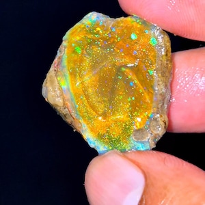 Könnte beinhalten: Ein roher, unpolierter Opal-Edelstein mit einer leuchtend gold-orangen Grundfarbe und intensiven grünen, blauen und gelben Schimmern auf der strukturierten Oberfläche. Der Stein hat eine unregelmäßige, organische Form mit rauen Kanten.
