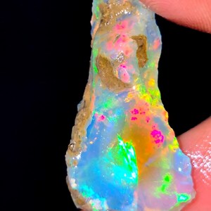 Könnte beinhalten: Ein roher, ungeschliffener Opal-Edelstein mit einem lebendigen Farbspiel aus Neonrosa, Elektroblau, leuchtendem Grün und Goldgelb auf einer durchscheinenden, milchig-weißen Basis, ergänzt durch natürliche, erdige braune Einschlüsse an der Seite.